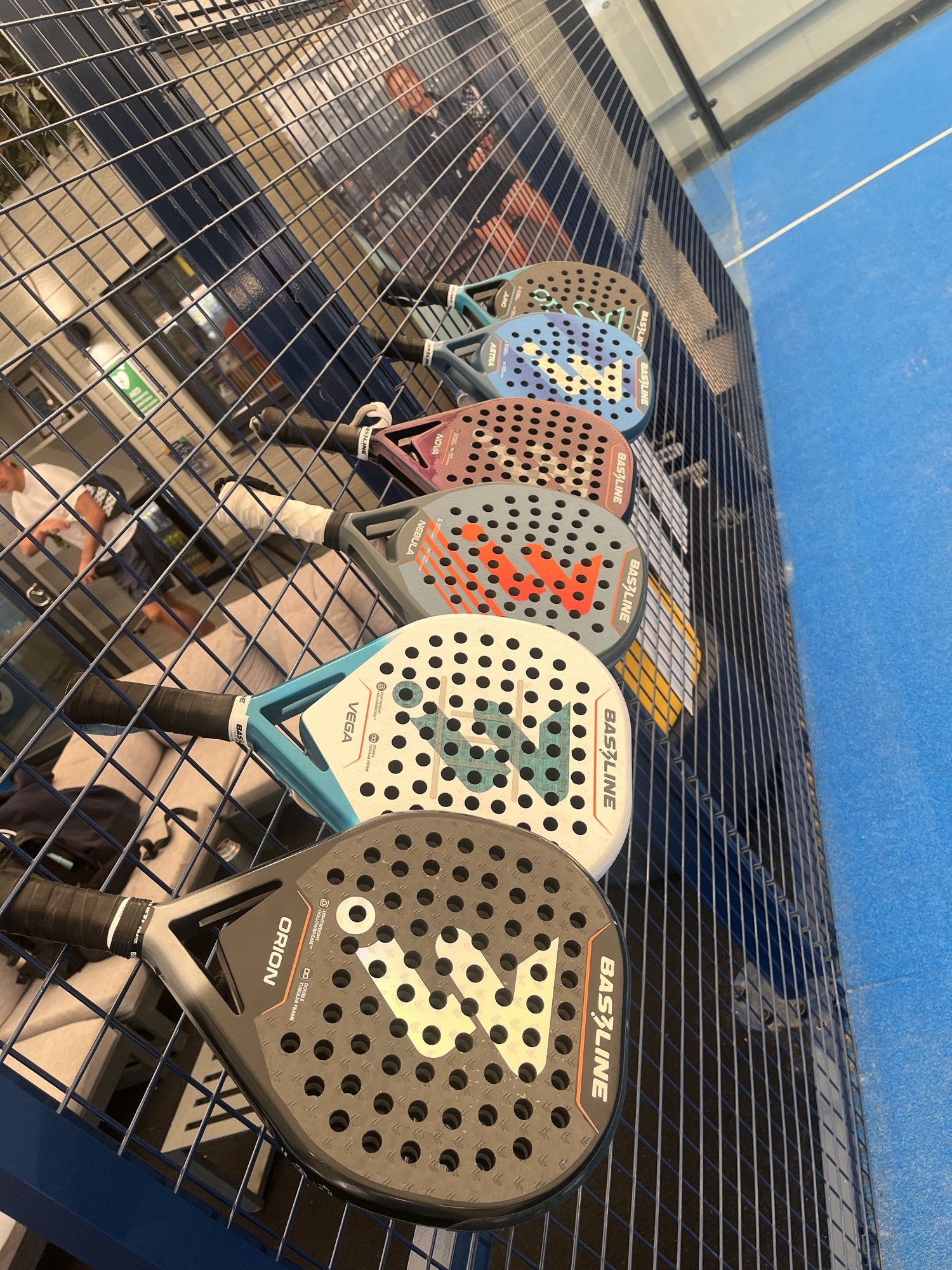 Six Bas3line teardrop rackets hung in a row on a court fence