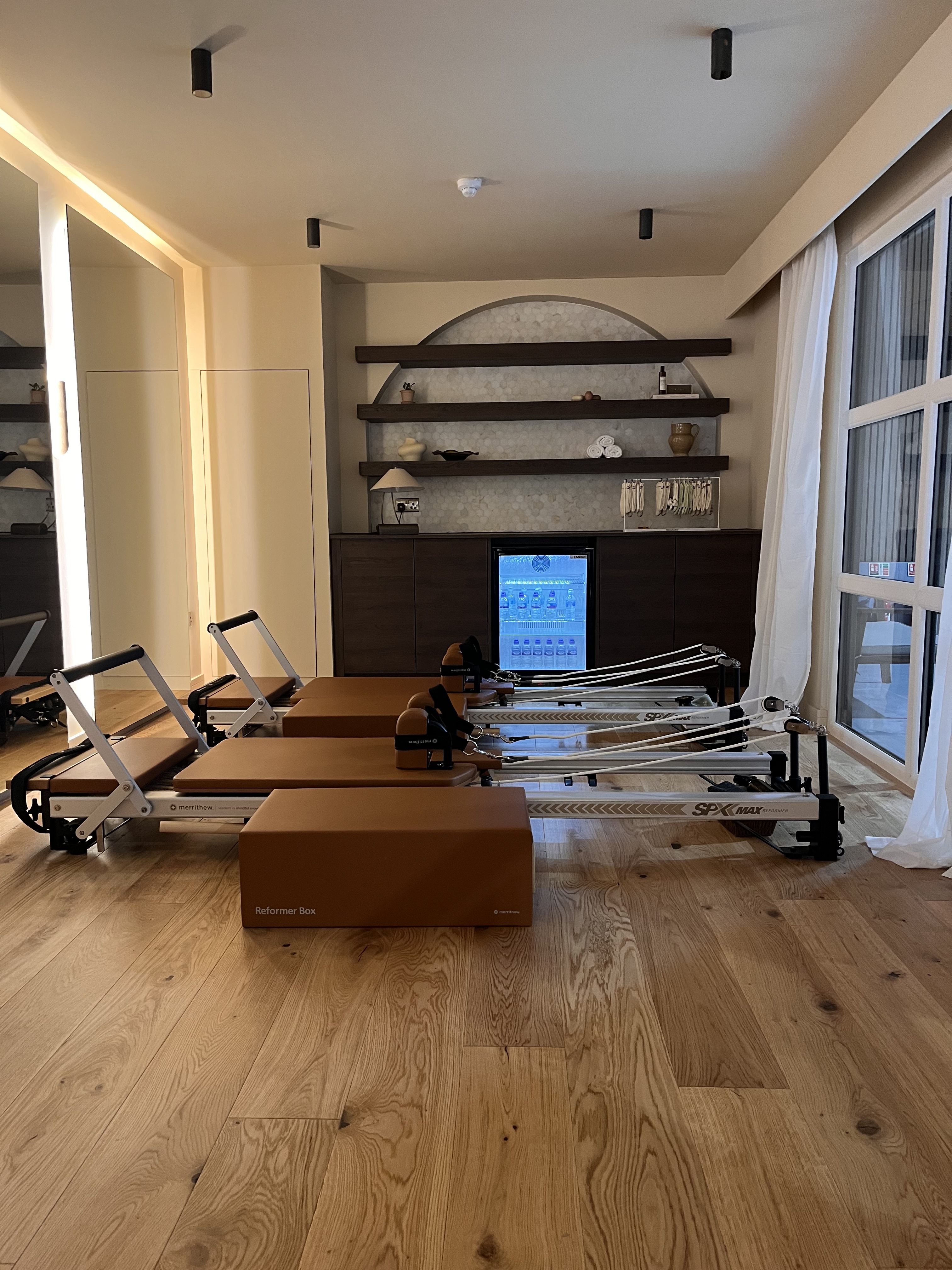 A row of Pilates reformers on an oak floor in a warmly lit studio with marble-tiled shelving behind