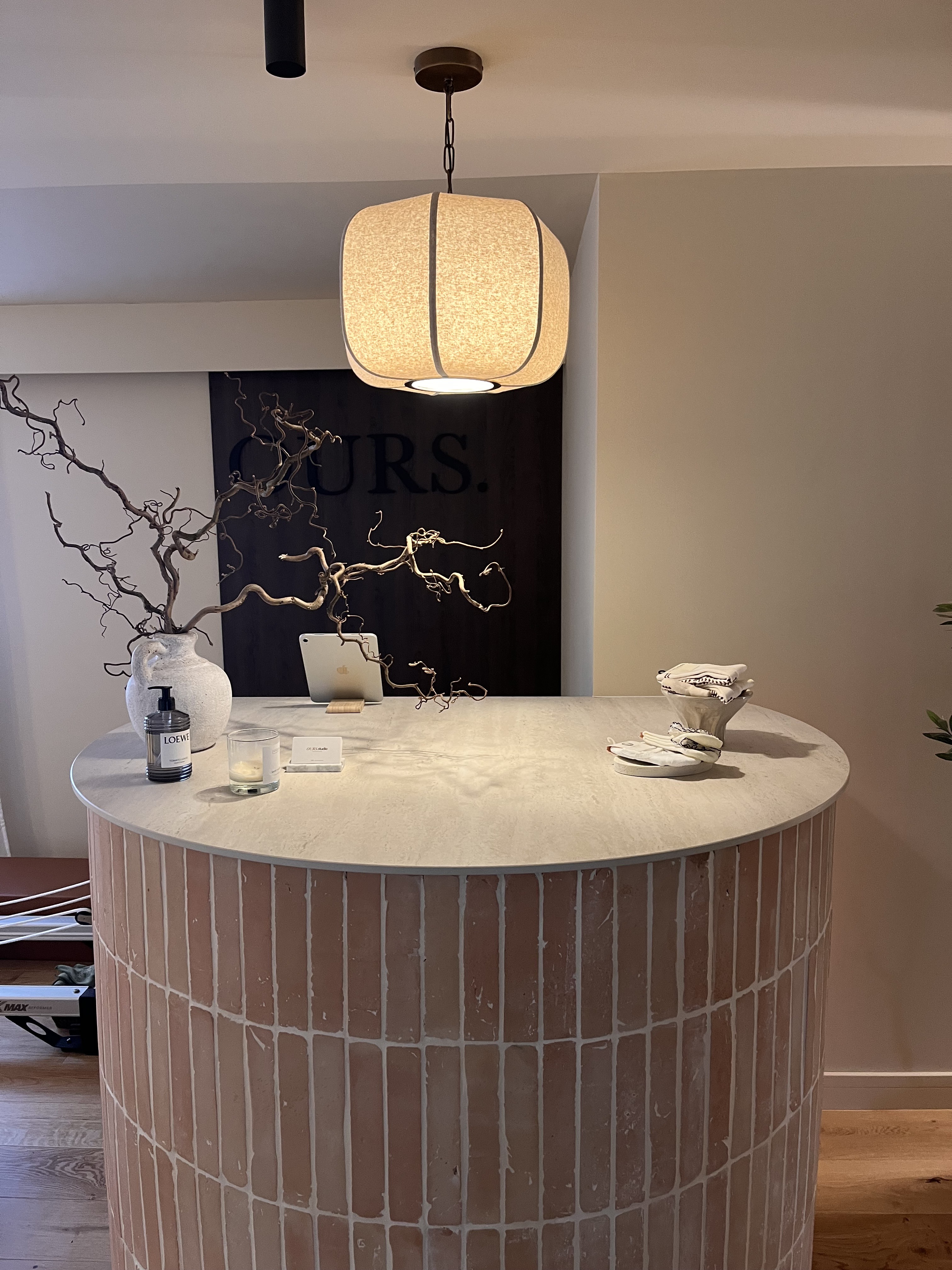 A curved terracotta-tiled studio counter with a ceramic jug of branches beneath the OURS wordmark