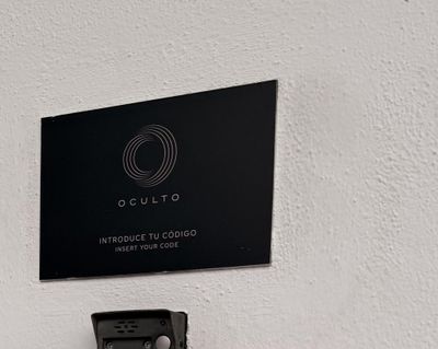 A black plaque on a white wall reading OCULTO, Introduce tu código, Insert your code