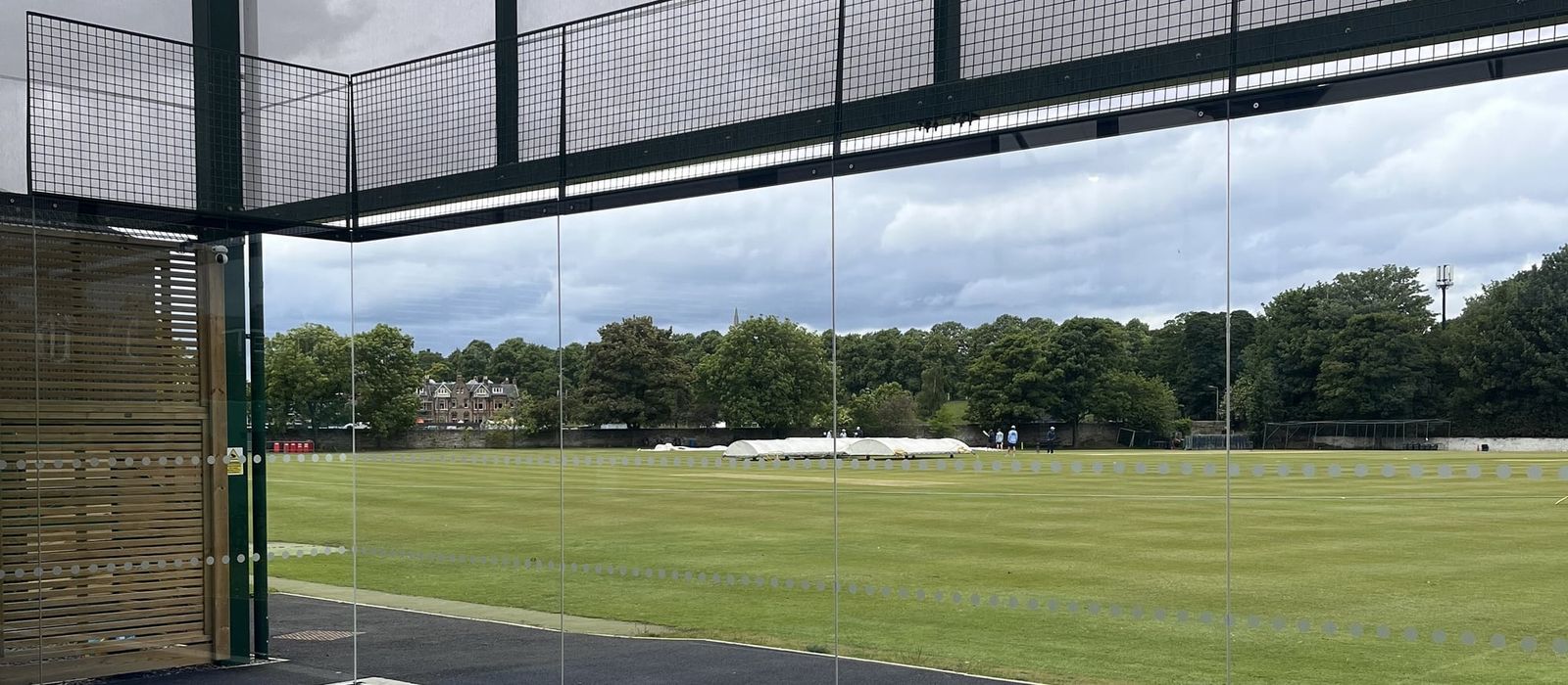 Looking out through the glass wall of a padel court, under its mesh-topped frame, to a cricket ground