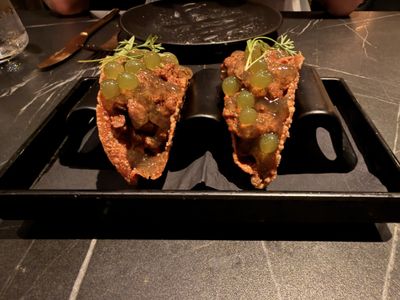 Two crisp tacos on a black tray, topped with green spheres that look like grapes