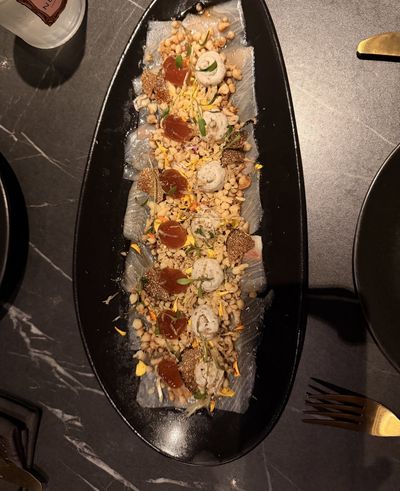 Thin slices of fish on a long black oval plate, dressed with truffle and tempura shavings